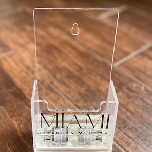 May include: A clear acrylic wall-mounted holder with a hole at the top for hanging. The holder has a white base and says "MIAMI WELLNESS MEDSPA" in black lettering. Two clear glass vials are inside the holder.