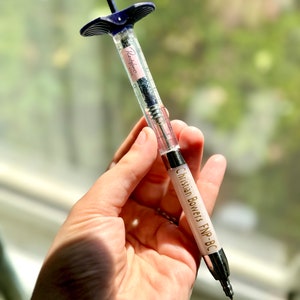 May include: A clear plastic syringe with a blue top and a white label that says "Restylane" and "0.5ml". The syringe is labeled with the text "Christian Bowers FNP-BC".