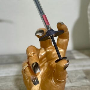 May include: A gold hand figurine with black and white nail polish and gold rings holds a clear syringe with a black cap and a black handle.