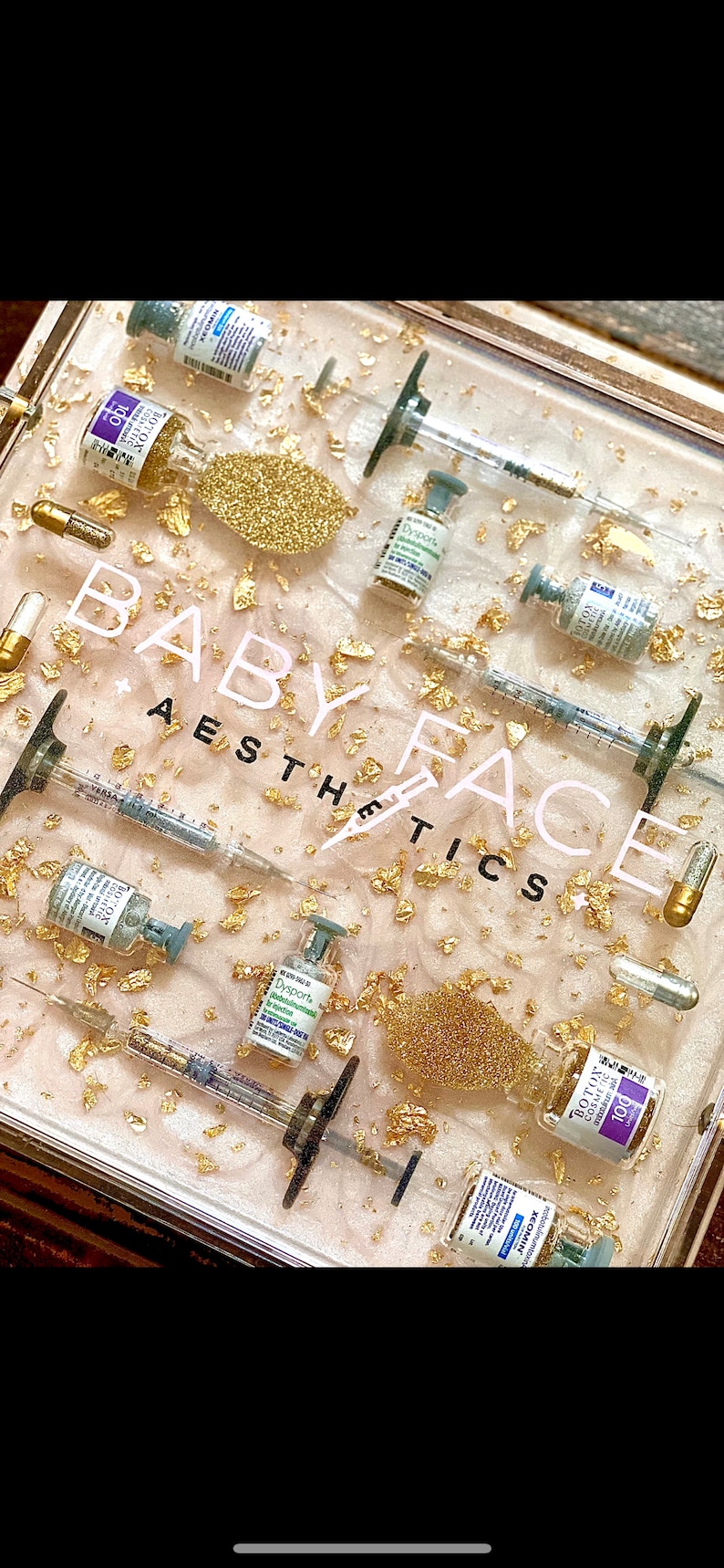 K&ouml;nnte beinhalten: Eine durchsichtige Plastikbox gef&uuml;llt mit goldenem Glitzer und kleinen Fl&auml;schchen mit Fl&uuml;ssigkeit. Die Fl&auml;schchen sind mit Namen wie "Botox" und "Dysport" beschriftet. Die Box tr&auml;gt au&szlig;erdem den Schriftzug "Baby Face Aesthetics".