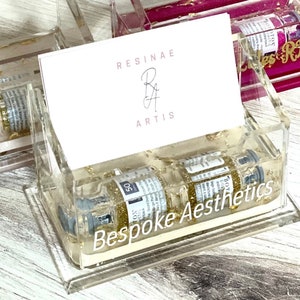 May include: A clear acrylic business card holder with gold glitter and two small glass vials filled with gold glitter. The card holder has the text "Resinae Artis" and "Bespoke Aesthetics" printed on it.