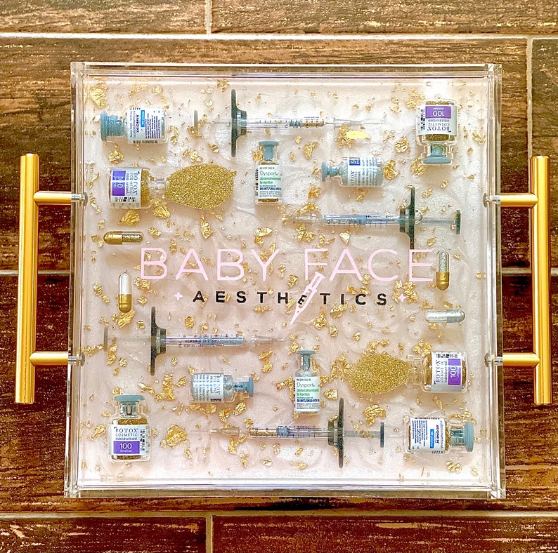 K&ouml;nnte beinhalten: Ein klares Acryltablett mit goldenen Flocken und verschiedenen Fl&auml;schchen und Spritzen. Das Tablett hat einen rosa Hintergrund mit dem Text "BABY FACE AESTHETICS" in Wei&szlig;.