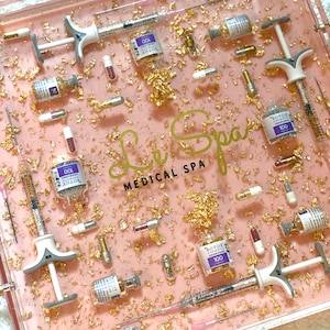K&ouml;nnte beinhalten: Ein rosa und klares Harztablett mit Goldflocken und silbernem Glitzer. Das Tablett enth&auml;lt eine Vielzahl von kleinen Fl&auml;schchen und Spritzen, darunter Fl&auml;schchen mit der Aufschrift "BOTOX COSMETIC 100" und "BOTOX 100". Das Tablett tr&auml;gt auch den Text "Le Spa MEDICAL SPA" in schwarzen Buchstaben.