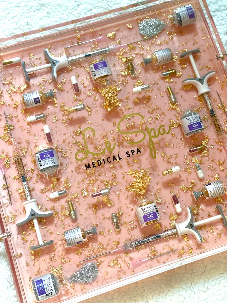 K&ouml;nnte beinhalten: Ein rosa und klares Harztablett mit Goldflocken und silbernem Glitzer. Das Tablett enth&auml;lt eine Vielzahl von kleinen Fl&auml;schchen und Spritzen, darunter Fl&auml;schchen mit der Aufschrift "BOTOX COSMETIC 100" und "BOTOX 100". Das Tablett tr&auml;gt auch den Text "Le Spa MEDICAL SPA" in schwarzen Buchstaben.