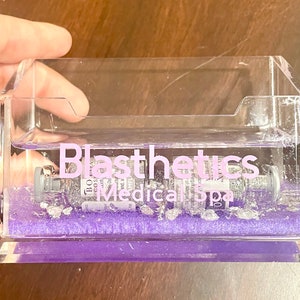May include: A clear acrylic business card holder with a purple base. The holder contains a small glass vial with a silver cap and the text "Blasthetics Medical Spa" in pink lettering.
