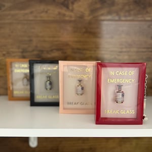 May include: Four framed miniature perfume displays in a row, each with a different colored frame: gold, black, peach, and red. Each frame has the text "IN CASE OF EMERGENCY BREAK GLASS" in gold lettering. A small perfume bottle is visible inside each frame.