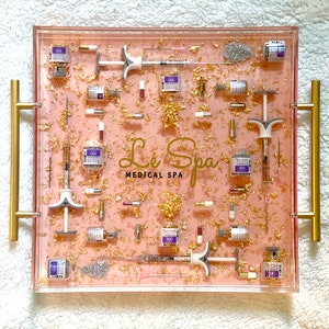 K&ouml;nnte beinhalten: Ein rosa Tablett mit Goldflocken und Glitzer, gef&uuml;llt mit Fl&auml;schchen und Spritzen. Das Tablett hat goldene Griffe und der Text "Le Spa Medical Spa" ist auf dem Tablett aufgedruckt.