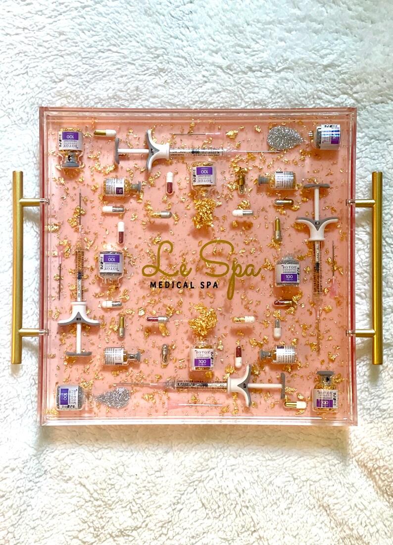 K&ouml;nnte beinhalten: Ein rosa Tablett mit Goldflocken und Glitzer, gef&uuml;llt mit Fl&auml;schchen und Spritzen. Das Tablett hat goldene Griffe und der Text "Le Spa Medical Spa" ist auf dem Tablett aufgedruckt.