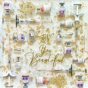 May include: A white tray with gold flakes and a variety of small glass vials, some with purple liquid and some with clear liquid. The vials are arranged in a grid pattern. The tray also has several syringes and the words "RN You Beautiful" written in gold lettering.