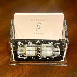 May include: A clear acrylic business card holder with a black base and silver flakes embedded in the resin. The holder has a pink business card inside with the text "RESINAE RA ARTIS".