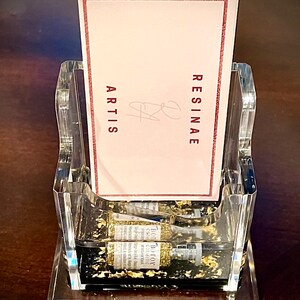 May include: A clear acrylic business card holder with a black base. The holder is filled with gold glitter and has two small bottles of gold glitter inside. The card holder has a pink card inside with the text "RESINAE ARTIS" in red.