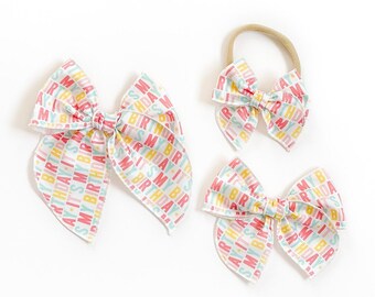 May include: Three fabric bows in various sizes, featuring a repeating "IT'S MY BIRTHDAY" pattern in pastel colors on a white background. One bow is attached to a nude-colored headband.