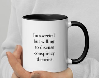 May include: White coffee mug with a black rim and handle. The mug has black text that reads "Introverted but willing to discuss conspiracy theories."