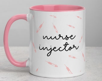 Może przedstawiać: Biały kubek ceramiczny z różowym rantem i uchwytem. Kubek ma wzór różowych strzykawek i napis "nurse injector" w kolorze czarnym.
