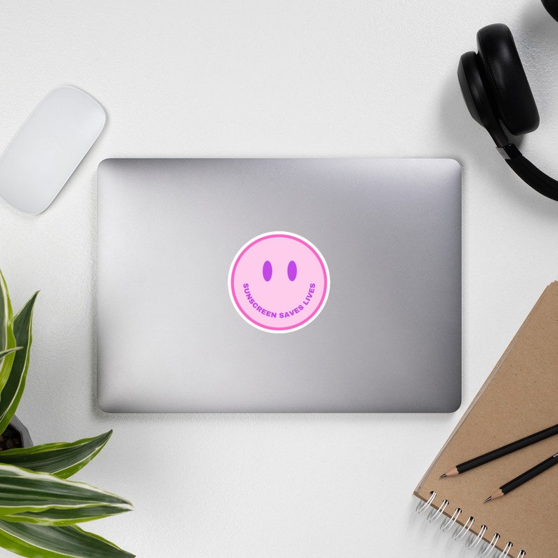 May include: A circular sticker featuring a light pink smiley face with two vertical purple oval eyes and a curved purple mouth. The text SUNSCREEN SAVES LIVES is printed in purple capital letters along the bottom curve of the smiley face.