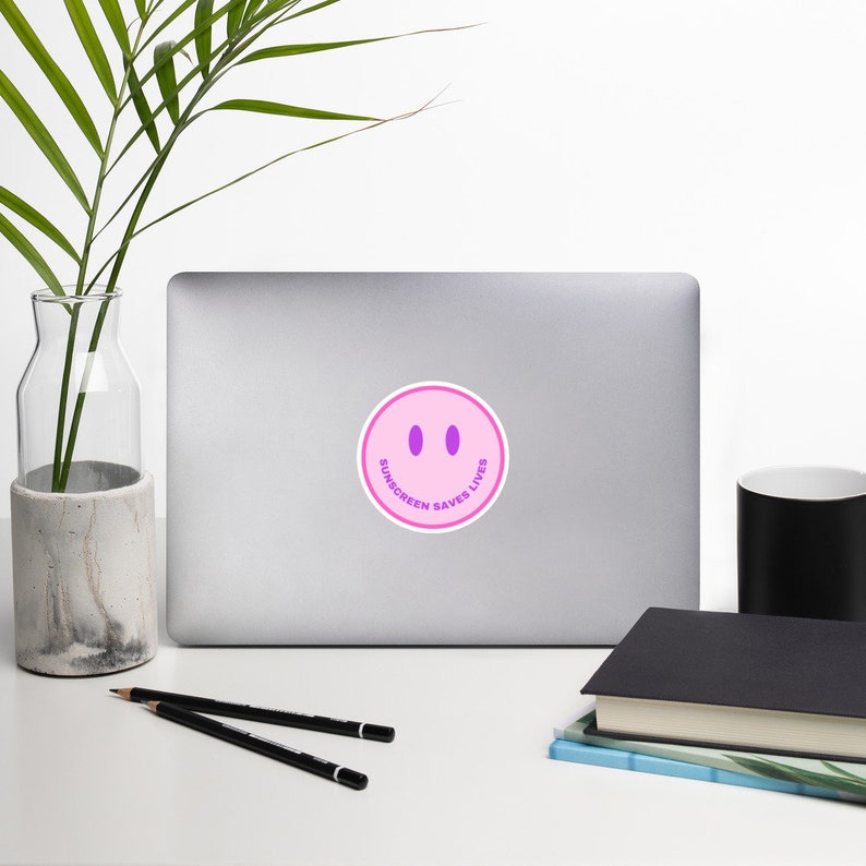 May include: A circular sticker featuring a light pink smiley face with two purple oval eyes and the text SUNSCREEN SAVES LIVES printed in purple along the bottom curve. The sticker is adhered to the center of a closed silver laptop computer.