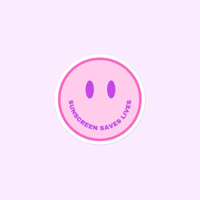 May include: A circular sticker featuring a light pink smiley face with two purple oval eyes. The text SUNSCREEN SAVES LIVES is printed in purple capital letters along the bottom curve of the face, framed by a bright pink circular border.