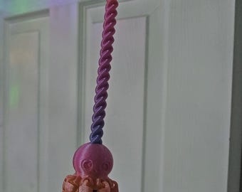 May include: A wand featuring a purple handle and a spiraled purple shaft topped with a pink octopus figure. The octopus has articulated, dangling tentacles in a lighter peach-pink hue. A small, dark blue star is attached to the very top of the wand.