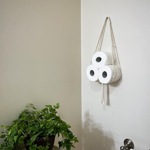 May include: Three rolls of toilet paper hanging from a macrame toilet paper holder with a white tassel. The holder is mounted on a white wall in a bathroom.