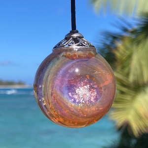 Pode incluir: Um pingente esf&eacute;rico de vidro com um design rodopiante e iridescente em tons de laranja, roxo e prata. O pingente &eacute; suspenso por um cord&atilde;o preto e tem uma tampa decorativa prateada. O fundo apresenta um c&eacute;u azul e o mar.