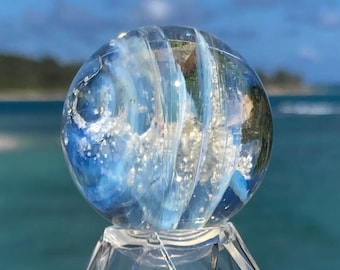 May include: A clear glass marble with blue and white swirls and glitter inside, displayed on a clear plastic stand.