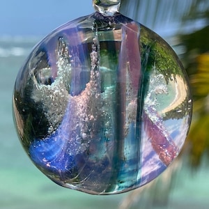 May include: A clear glass pendant with a spherical shape, displaying a blend of colors including blue, pink, and green. The pendant is suspended by a black cord, with the ocean and palm trees in the background.