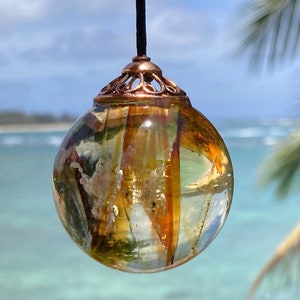 Könnte beinhalten: Ein runder, transparenter Glasanhänger mit einem kupferfarbenen Metalldeckel. Das Glas enthält verschiedene Brauntöne, Gelb- und Weißtöne, die an natürliche Elemente erinnern. Der Anhänger hängt vor einem verschwommenen Hintergrund aus blauem Wasser und einer Palme.