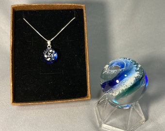 May include: A silver necklace with a blue and white glass pendant in a black box. A blue and white glass orb on a clear stand.