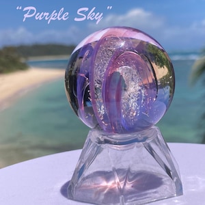 Pode incluir: Uma esfera de vidro transparente com redemoinhos roxos e prateados, repousando sobre uma base de vidro transparente facetada. A esfera est&aacute; rotulada como "Purple Sky" em uma fonte cursiva. O fundo mostra uma praia e o oceano.