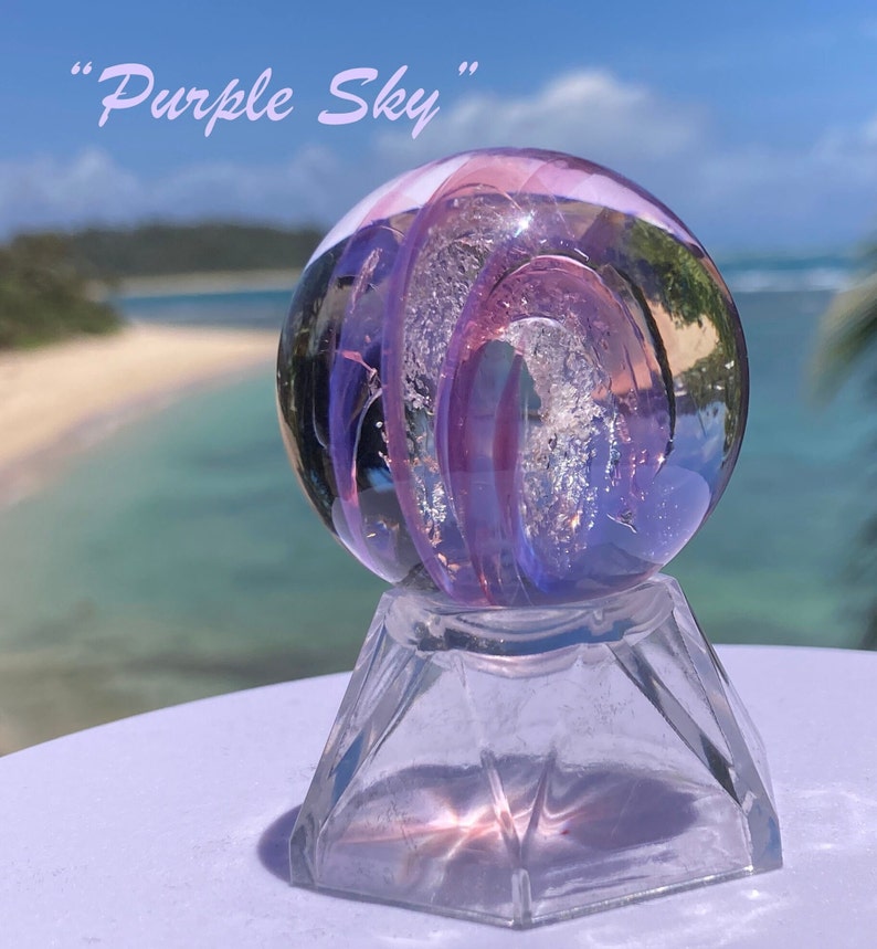 Pode incluir: Uma esfera de vidro transparente com redemoinhos roxos e prateados, repousando sobre uma base de vidro transparente facetada. A esfera est&aacute; rotulada como "Purple Sky" em uma fonte cursiva. O fundo mostra uma praia e o oceano.