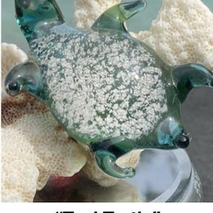 Pode incluir: Uma estatueta de tartaruga de vidro verde-azulado com uma carapa&ccedil;a transparente. A tartaruga tem um design detalhado com barbatanas, cabe&ccedil;a e cauda. A carapa&ccedil;a tem um padr&atilde;o salpicado branco. A estatueta &eacute; exibida em uma superf&iacute;cie branca semelhante a coral.