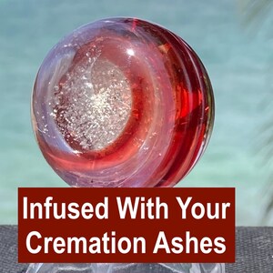 Pode incluir: Um globo de vidro transparente com redemoinhos vermelhos e roxos, infundido com cinzas de crema&ccedil;&atilde;o. O globo tem um n&uacute;cleo central prateado. O texto em fundo vermelho diz "Infused With Your Cremation Ashes."
