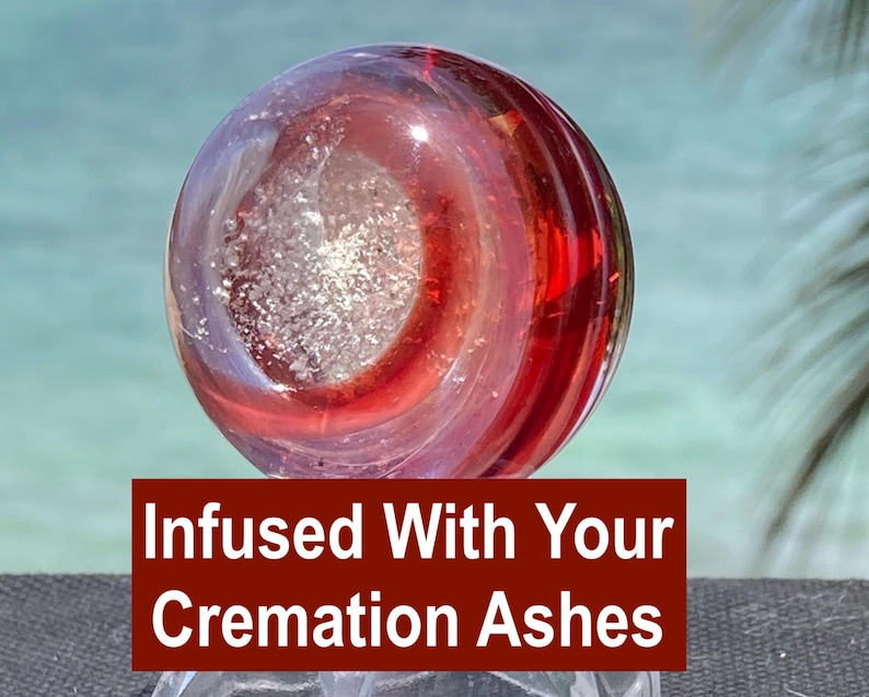 Pode incluir: Um globo de vidro transparente com redemoinhos vermelhos e roxos, infundido com cinzas de crema&ccedil;&atilde;o. O globo tem um n&uacute;cleo central prateado. O texto em fundo vermelho diz "Infused With Your Cremation Ashes."