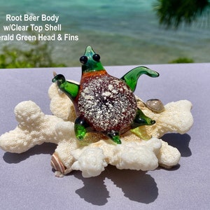 May include: A glass sea turtle figurine with a root beer colored body, clear top shell, and emerald green head and fins. The turtle is sitting on a white coral base.