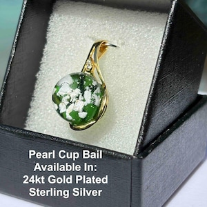 Pode incluir: Um pingente banhado a ouro com um globo de vidro verde e branco, apresentado em uma caixa de presente preta. O pingente tem um design curvo com um pequeno detalhe de diamante. O texto na caixa diz "Pearl Cup Bail Available In: 24kt Gold Plated Sterling Silver."