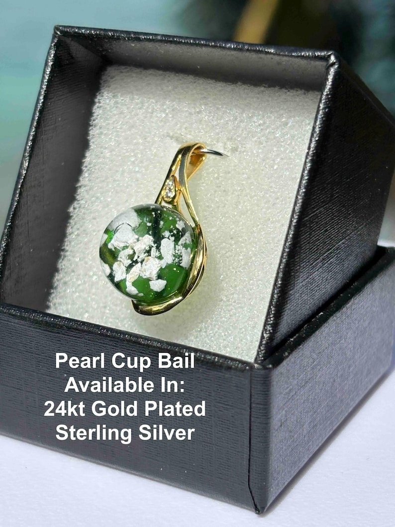 Pode incluir: Um pingente banhado a ouro com um globo de vidro verde e branco, apresentado em uma caixa de presente preta. O pingente tem um design curvo com um pequeno detalhe de diamante. O texto na caixa diz "Pearl Cup Bail Available In: 24kt Gold Plated Sterling Silver."