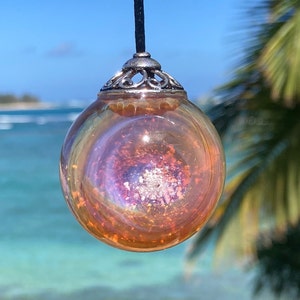 Pode incluir: Um pingente esf&eacute;rico transparente com um interior iridescente e rodopiante de laranja, roxo e prata. O pingente &eacute; suspenso por um cord&atilde;o preto e tem uma tampa decorativa prateada. O fundo apresenta uma cena de praia tropical.