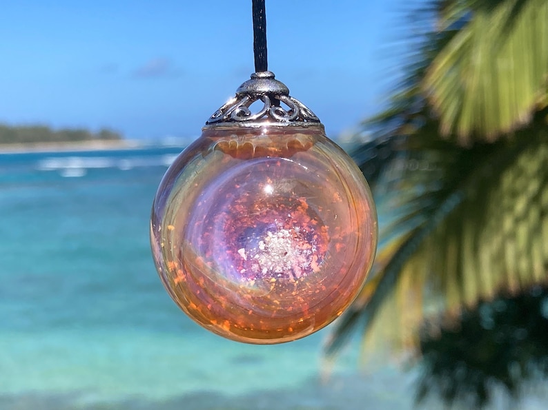 Pode incluir: Um pingente esf&eacute;rico transparente com um interior iridescente e rodopiante de laranja, roxo e prata. O pingente &eacute; suspenso por um cord&atilde;o preto e tem uma tampa decorativa prateada. O fundo apresenta uma cena de praia tropical.