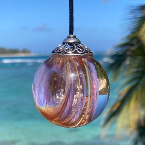 Pode incluir: Um pendente redondo de vidro iridescente com um padr&atilde;o rodopiante de tons laranja, roxo e dourado. O pendente &eacute; suspenso por um cord&atilde;o preto e tem uma tampa decorativa de metal prateado. O fundo apresenta uma vista desfocada do mar e do c&eacute;u.