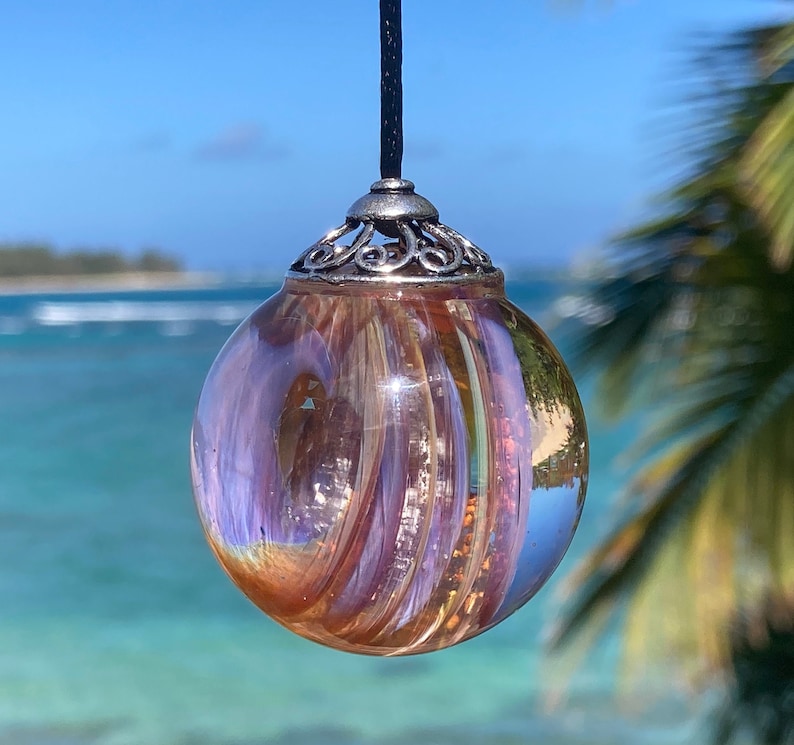 Pode incluir: Um pendente redondo de vidro iridescente com um padr&atilde;o rodopiante de tons laranja, roxo e dourado. O pendente &eacute; suspenso por um cord&atilde;o preto e tem uma tampa decorativa de metal prateado. O fundo apresenta uma vista desfocada do mar e do c&eacute;u.