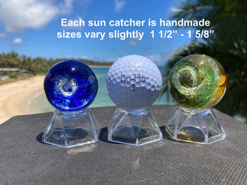 Pode incluir: Tr&ecirc;s apanhadores de sol feitos &agrave; m&atilde;o em suportes transparentes. Os apanhadores de sol incluem um globo de vidro azul, uma bola de golfe branca e um globo de vidro verde e amarelo. O texto diz: "Cada apanhador de sol &eacute; feito &agrave; m&atilde;o, os tamanhos variam ligeiramente 3,8 cm - 4,1 cm".
