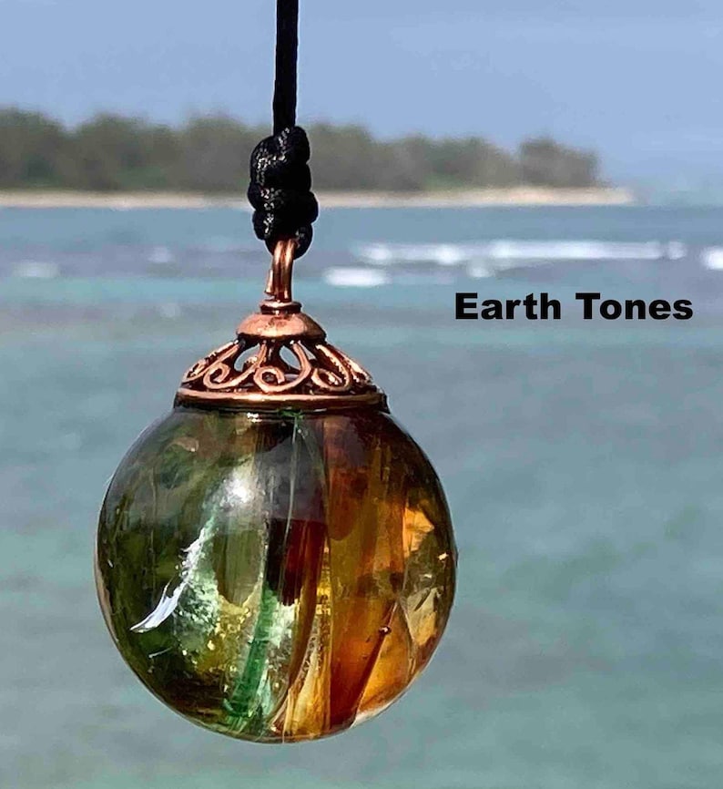 Pode incluir: Um pingente esf&eacute;rico transparente com tons verdes e &acirc;mbar, suspenso por um cord&atilde;o preto. O pingente tem uma tampa decorativa cor de cobre. O texto "Earth Tones" &eacute; vis&iacute;vel na imagem.