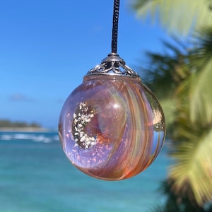 Pode incluir: Um pingente esf&eacute;rico transparente com uma tampa prateada e um cord&atilde;o preto. O pingente tem um design girat&oacute;rio e iridescente em tons de laranja, roxo e dourado, com detalhes brancos embutidos. O fundo mostra um c&eacute;u azul e o oceano.