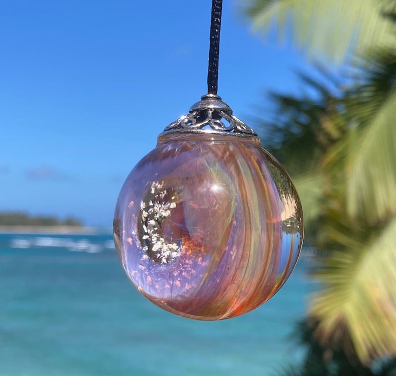 Pode incluir: Um pingente esf&eacute;rico transparente com uma tampa prateada e um cord&atilde;o preto. O pingente tem um design girat&oacute;rio e iridescente em tons de laranja, roxo e dourado, com detalhes brancos embutidos. O fundo mostra um c&eacute;u azul e o oceano.