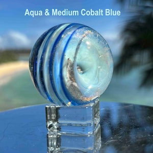 Pode incluir: Uma bolinha de gude de vidro transparente com redemoinhos azul-&aacute;gua e azul cobalto m&eacute;dio, repousando sobre um cubo de vidro transparente. A bolinha de gude tem um design em espiral e o texto "Aqua & Medium Cobalt Blue" &eacute; vis&iacute;vel.