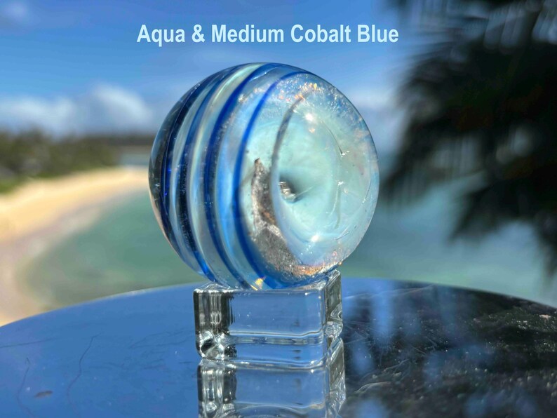 Pode incluir: Uma bolinha de gude de vidro transparente com redemoinhos azul-&aacute;gua e azul cobalto m&eacute;dio, repousando sobre um cubo de vidro transparente. A bolinha de gude tem um design em espiral e o texto "Aqua & Medium Cobalt Blue" &eacute; vis&iacute;vel.