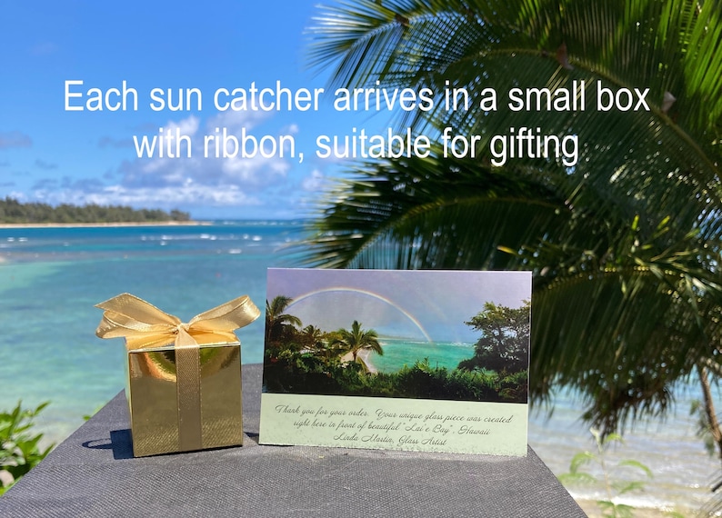 Pode incluir: Uma caixa de presente dourada com um la&ccedil;o de fita a condizer, um cart&atilde;o com um arco-&iacute;ris sobre o oceano e o texto "Each sun catcher arrives in a small box with ribbon, suitable for gifting."