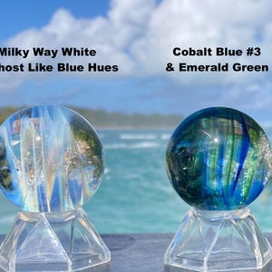 Pode incluir: Duas esferas de vidro em suportes transparentes. Uma esfera &eacute; branca leitosa com tons azuis, a outra &eacute; azul cobalto e verde esmeralda. O texto na imagem diz "Milky Way White w/Ghost Like Blue Hues" e "Cobalt Blue #3 & Emerald Green."