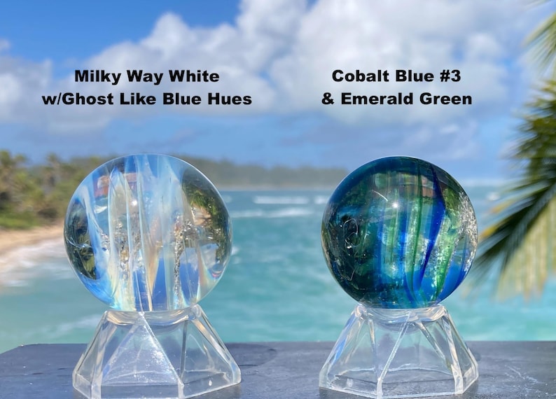 Pode incluir: Duas esferas de vidro em suportes transparentes. Uma esfera &eacute; branca leitosa com tons azuis, a outra &eacute; azul cobalto e verde esmeralda. O texto na imagem diz "Milky Way White w/Ghost Like Blue Hues" e "Cobalt Blue #3 & Emerald Green."