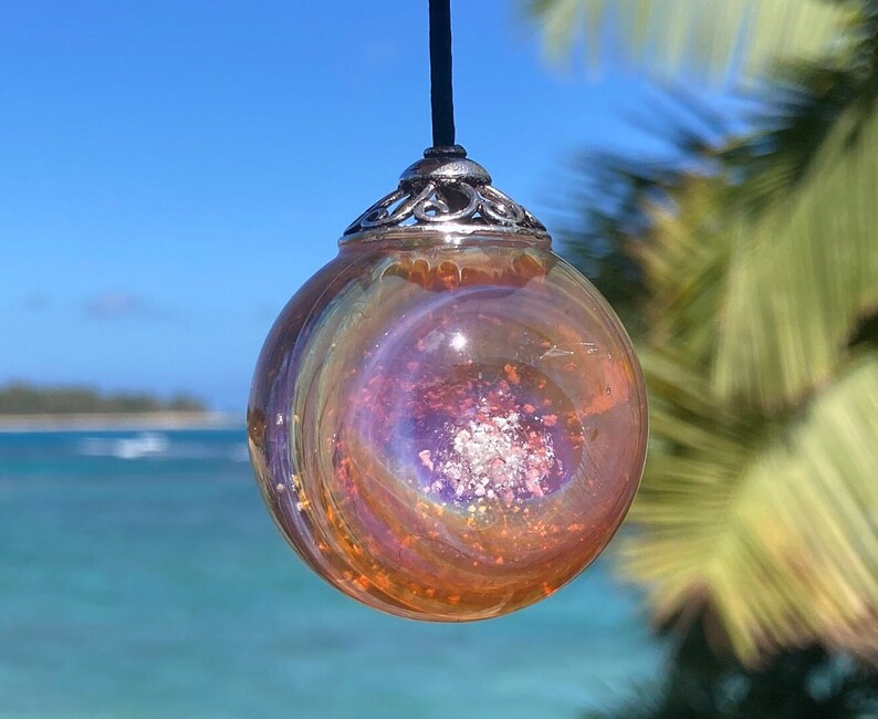 Pode incluir: Um pingente esf&eacute;rico de vidro com um design rodopiante e iridescente em tons de laranja, roxo e prata. O pingente &eacute; suspenso por um cord&atilde;o preto e tem uma tampa decorativa prateada. O fundo apresenta um c&eacute;u azul e o mar.